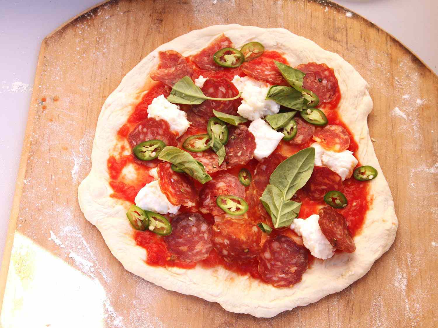 Overhead shot of pizza dough rolled out, topped with tomato sauce, sliced spicy salami, mozzarella, sliced chiles, and fresh basil leaves 
