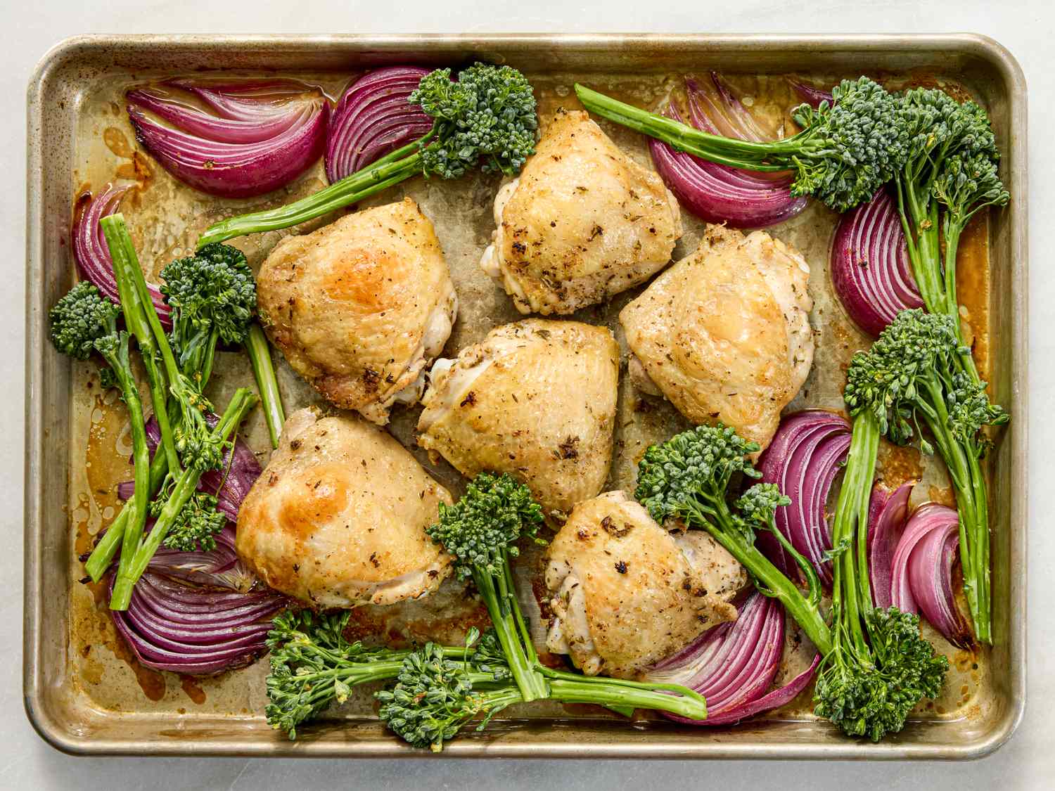 Sheet pan with roasted chicken thighs red onion slices and broccolini spears
