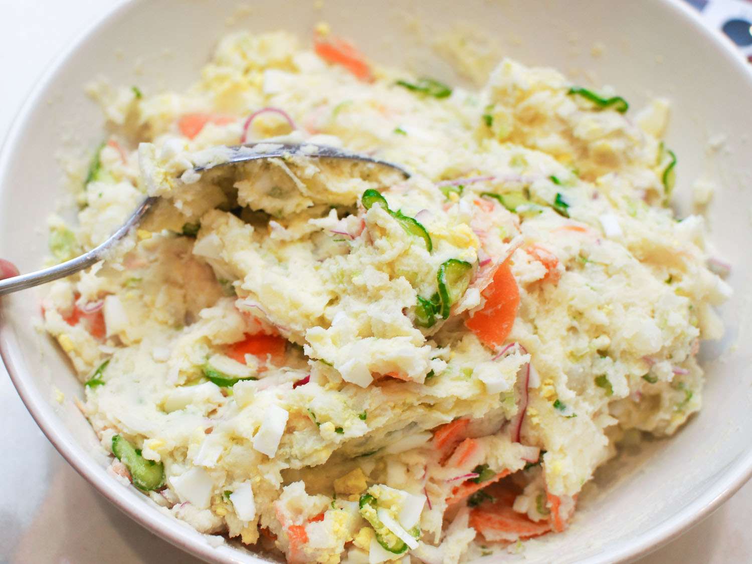 The potato salad, fully assembled in a mixing bowl, with a slotted spoon scooping into the mixture.