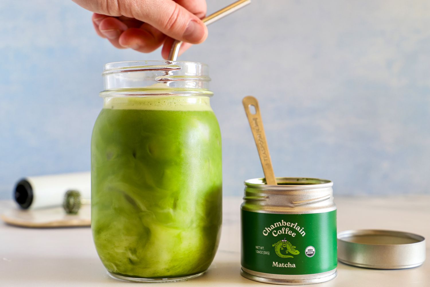 A person mixing a matcha latte in a glass with a tin of matcha powders beside it.