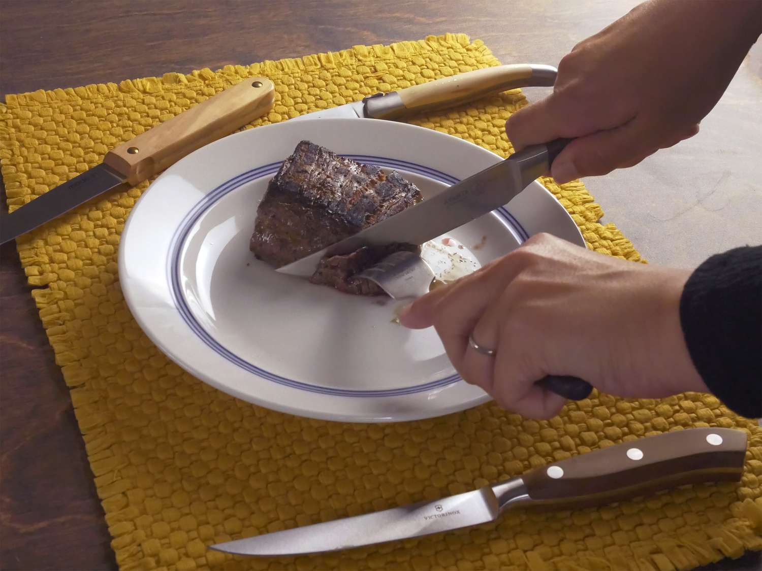 Hands using a steak knife to cut meat on a plate with other steak knives on placemat