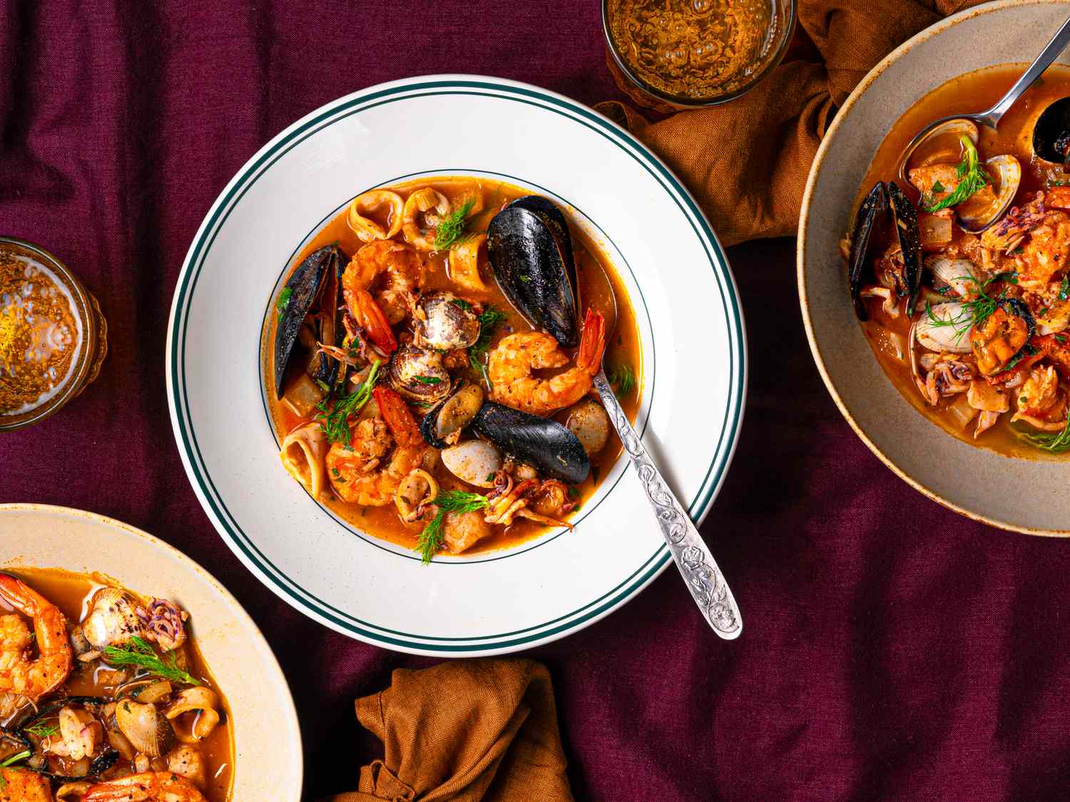 Three bowls of seafood stew garnished with green herbs served on a purple cloth with glasses of drinks
