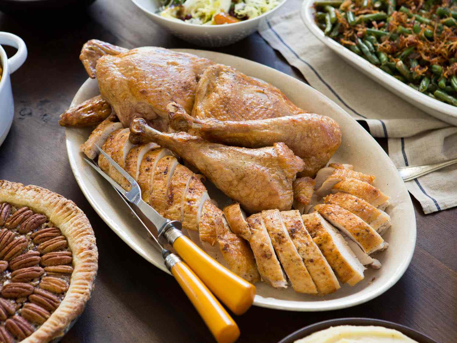 A platter of carved turkey with a carving knife and fork, surrounded by pecan pie, green bean casserole, and salad.