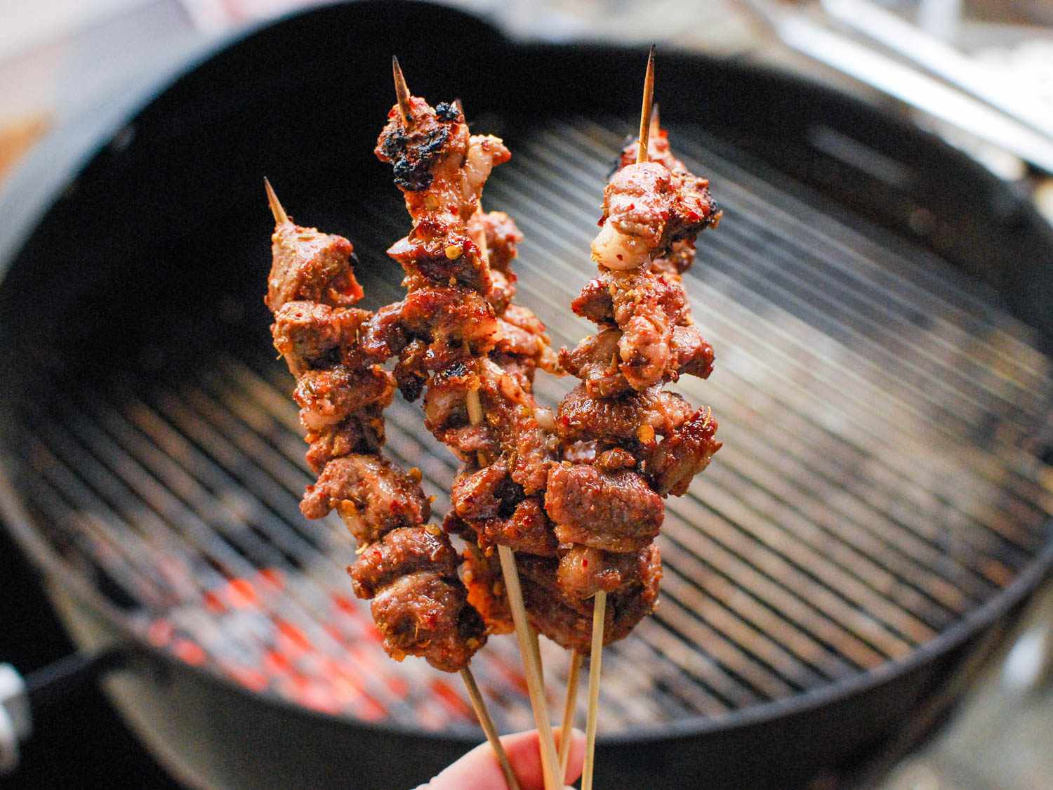 Holding four cooked lamb skewers with a grill in the background.