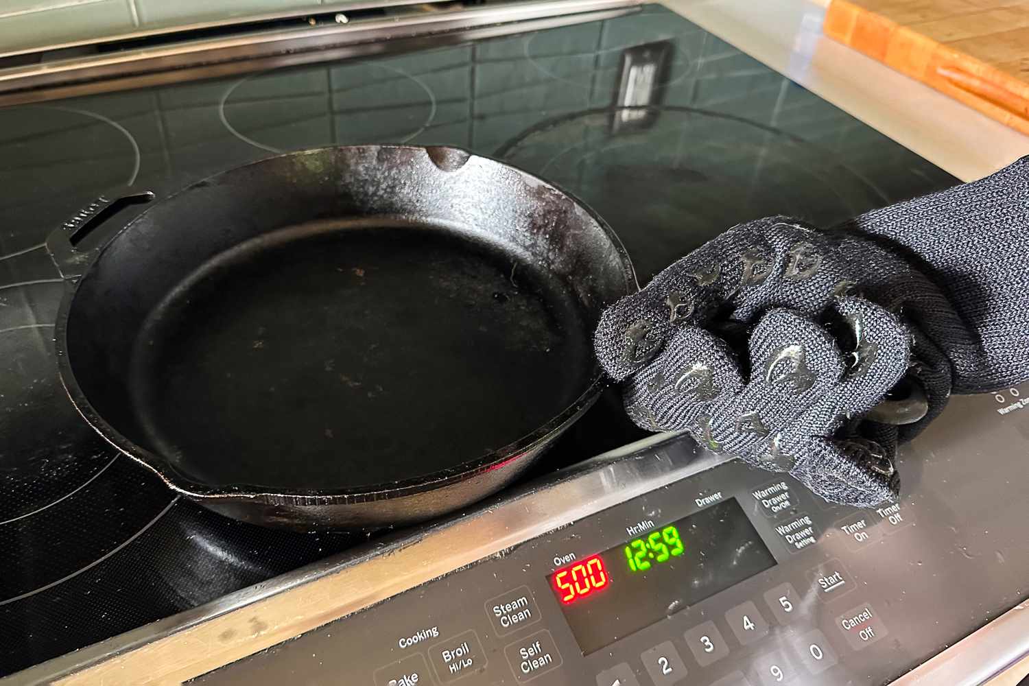 A cast iron skillet on a stove top with a gloved hand holding it oven temperature and time displayed