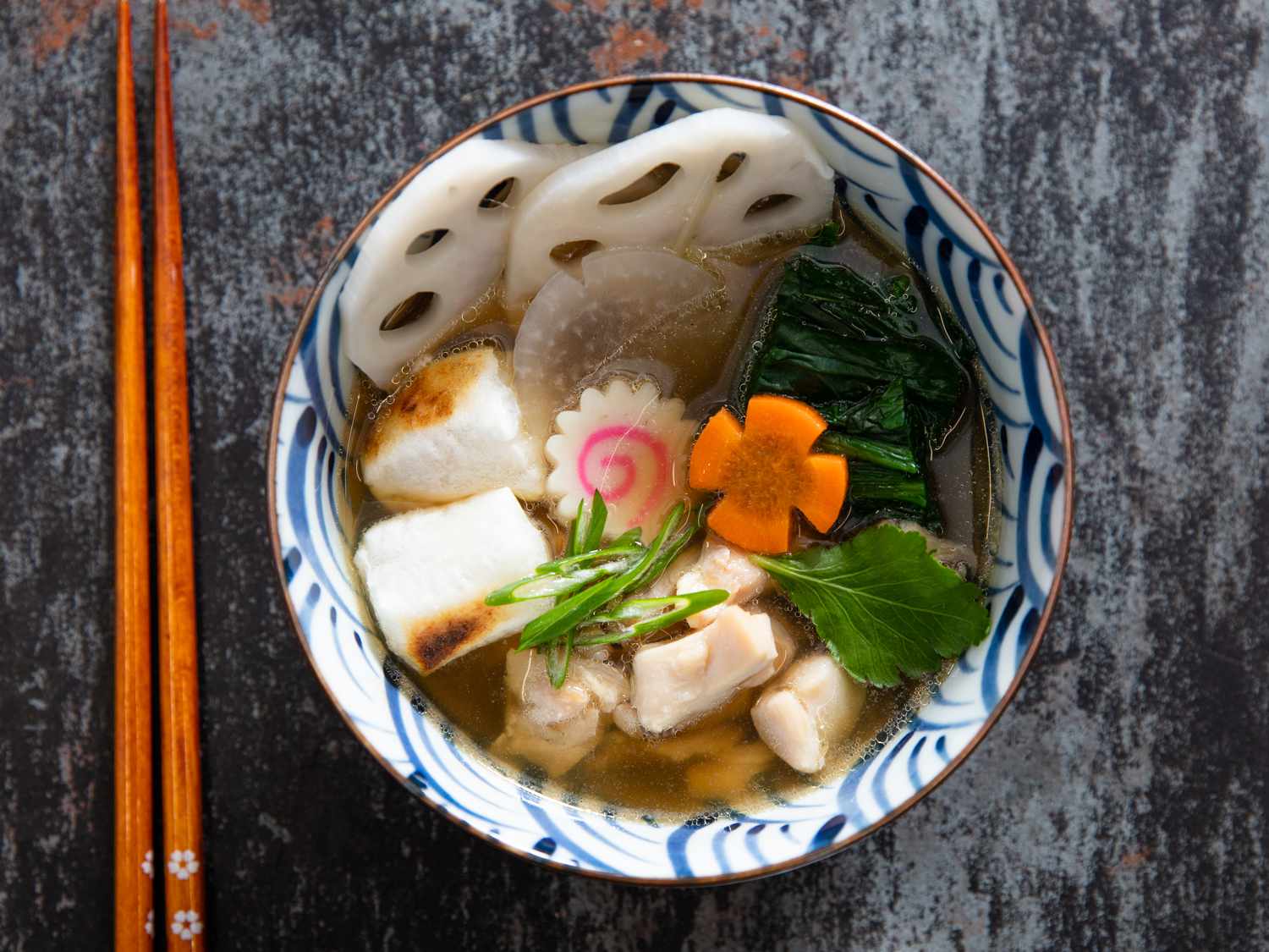 A bowl of Japanese ozoni soup with sliced lotus root, chicken, mochi, and vegetables next to a pair of chopsticks.