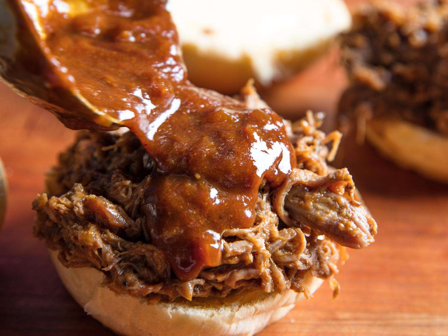Closeup of a pulled lamb piled on the bottom half of a bun getting topped with more sauce.