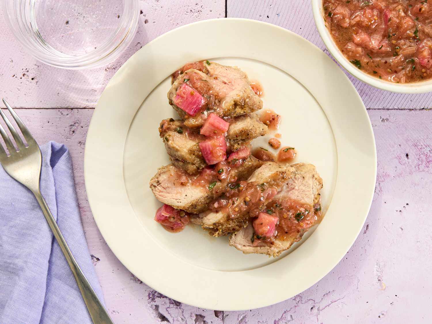 sliced pork with rhubarb sauce drizzled on it, on aa cream colored plate, with bowl of rhubarb sauce to the side, fork and napkin, on a tile surface.