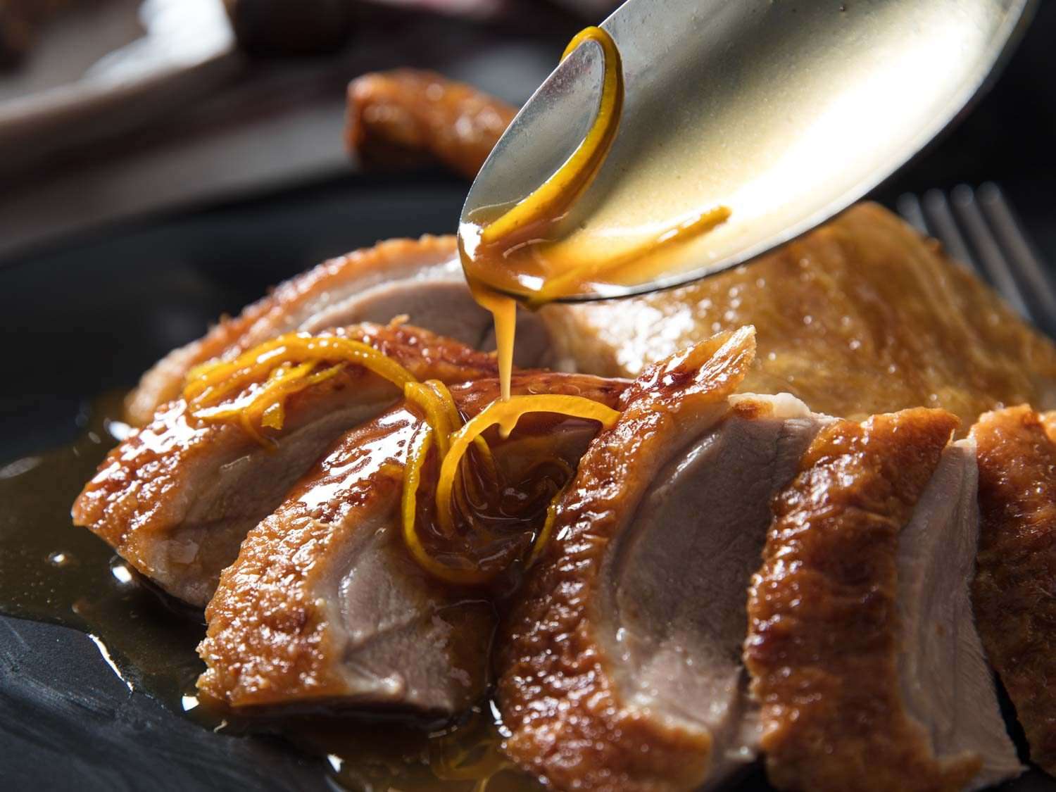 A spoonful of sauce and orange zest being poured onto sliced roast duck breast for Duck à l'Orange