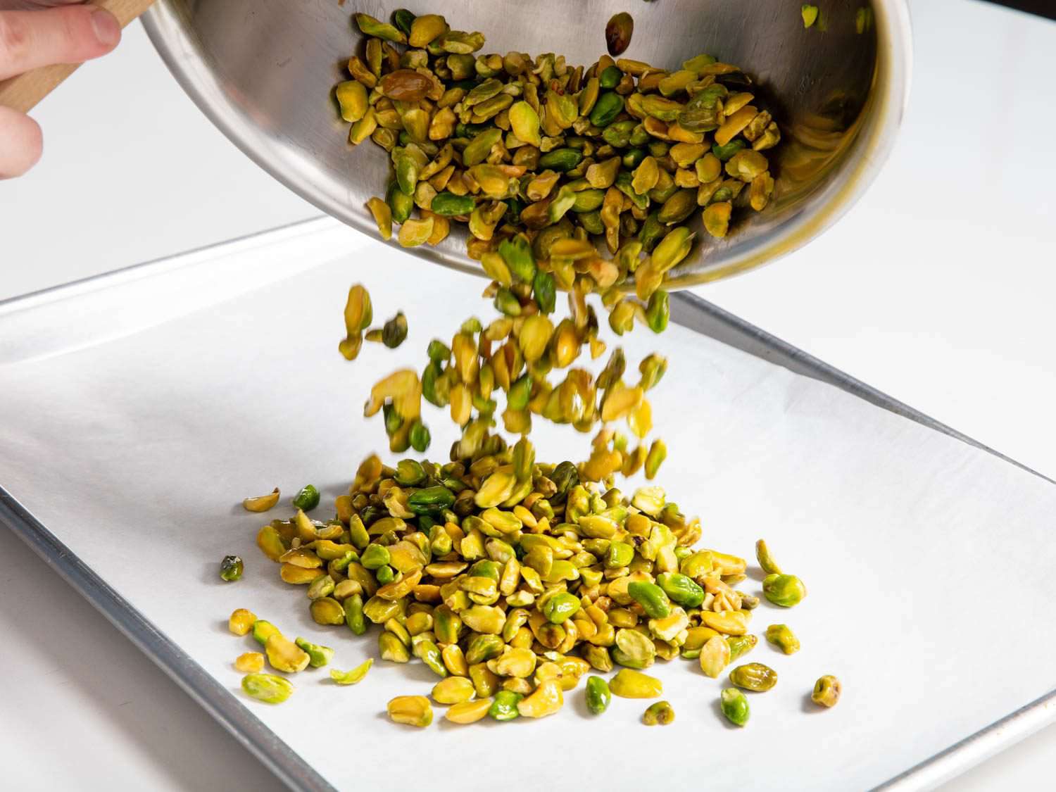 Blanched, peeled, and oiled pistachios being transferred to a parchment-lined sheet pan.