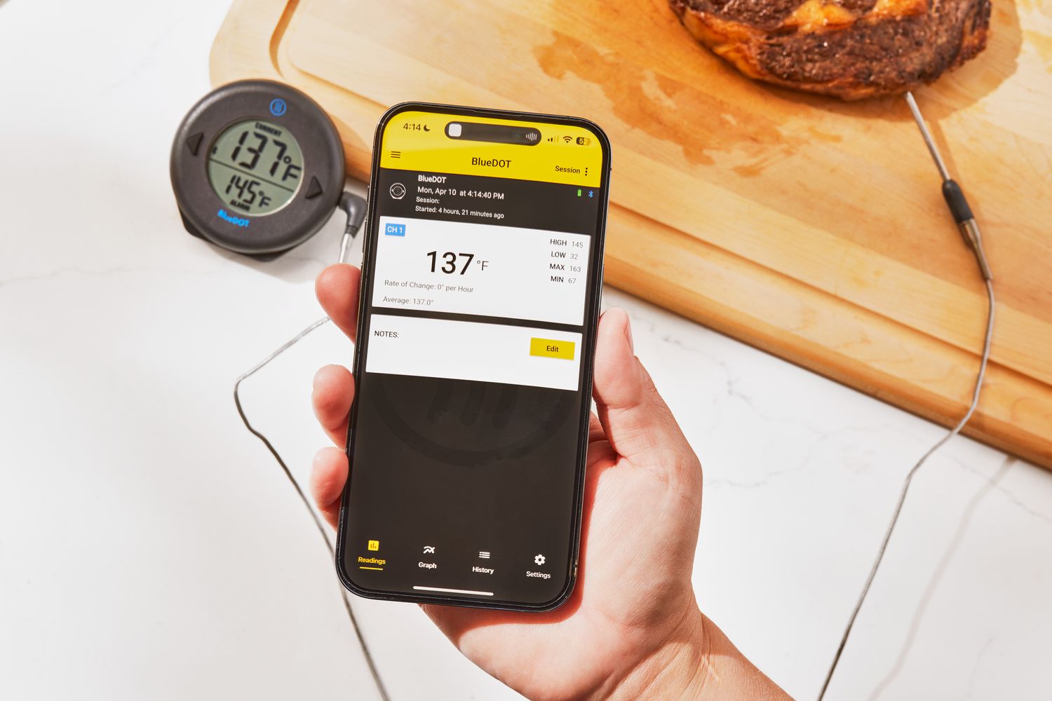 a hand holding a phone and showing a thermometer's app as meat rest on a cutting board in the background