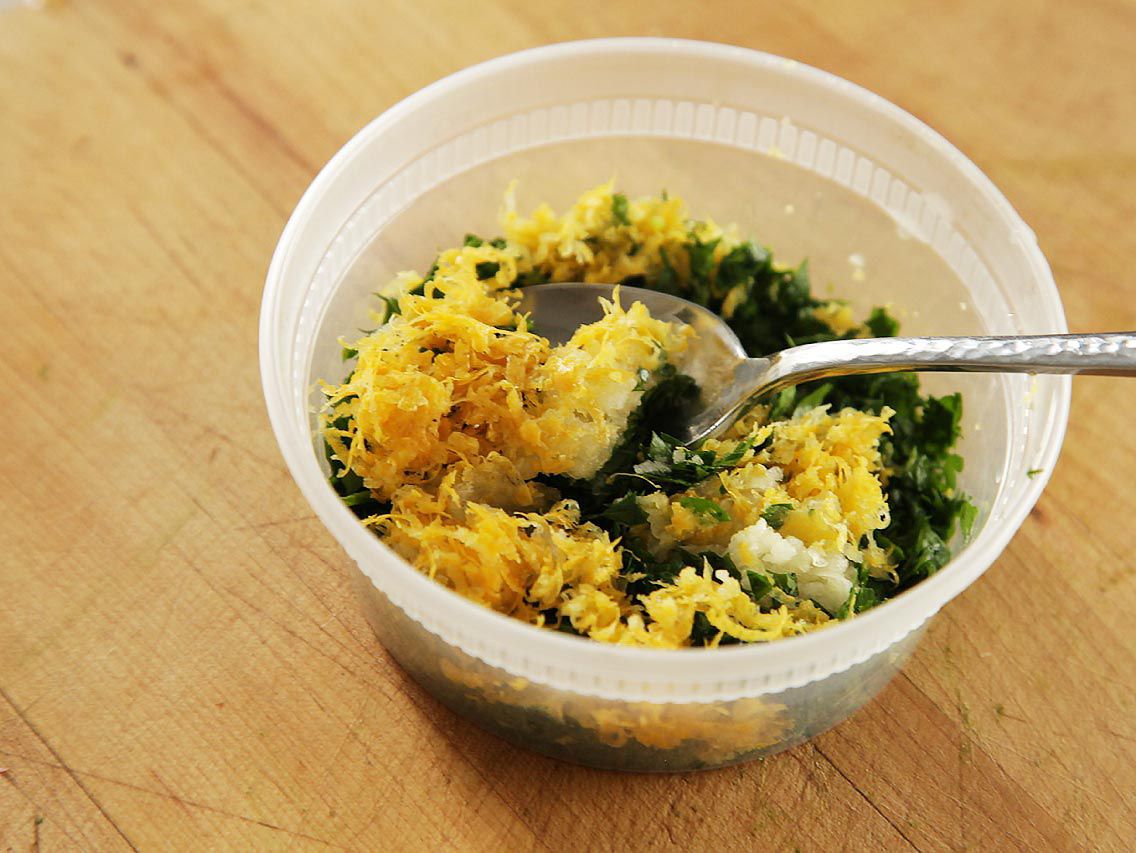 Plastic container filled with lemon zest, chopped herbs, and garlic with a spoon in it.
