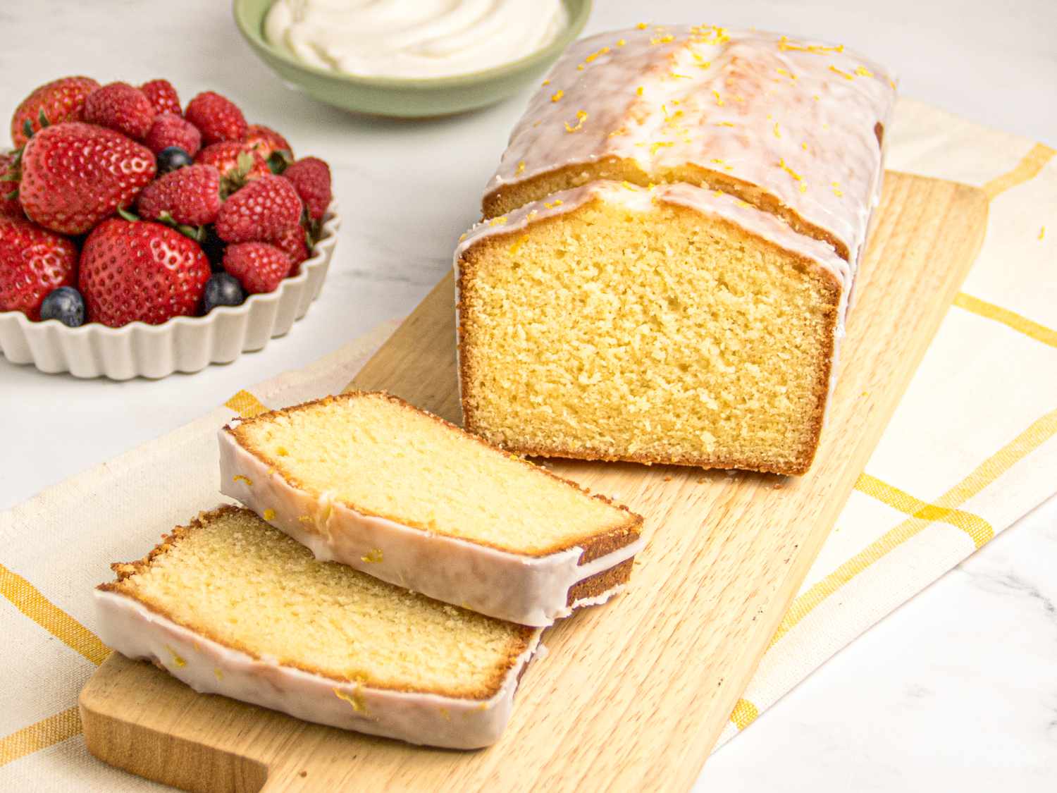 Lemon pound cake with icing, two slices cut, served on a wooden board, bowl of mixed berries and whipped cream in the background.