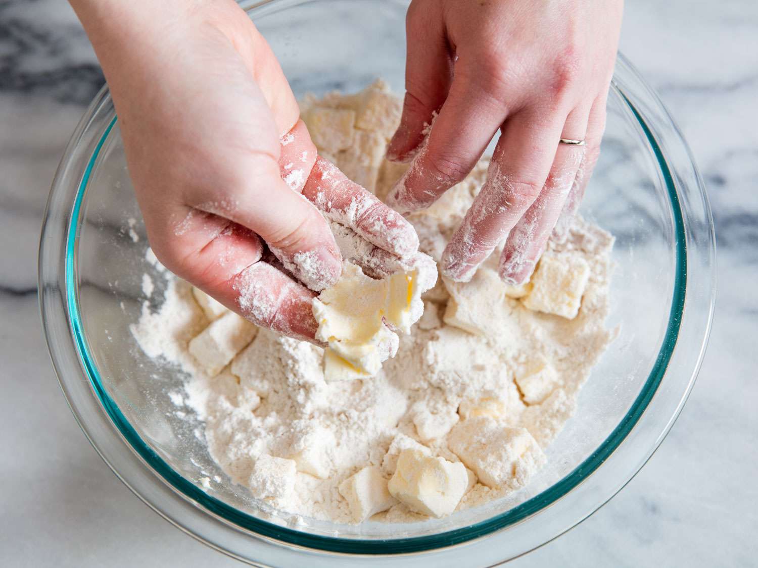 Hands working with a glass bowl of flour and butter, smashing flour-coated butter cubes flat