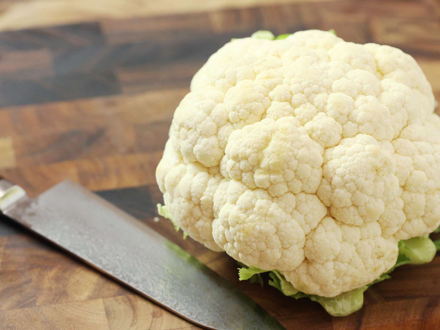 A head of cauliflower sitting next to a chef's knife on a wooden board