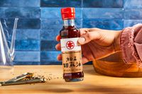 A hand holding a bottle of pure sesame seed oil labeled with Japanese text, on a wooden countertop with teaspoons and black sesame seeds