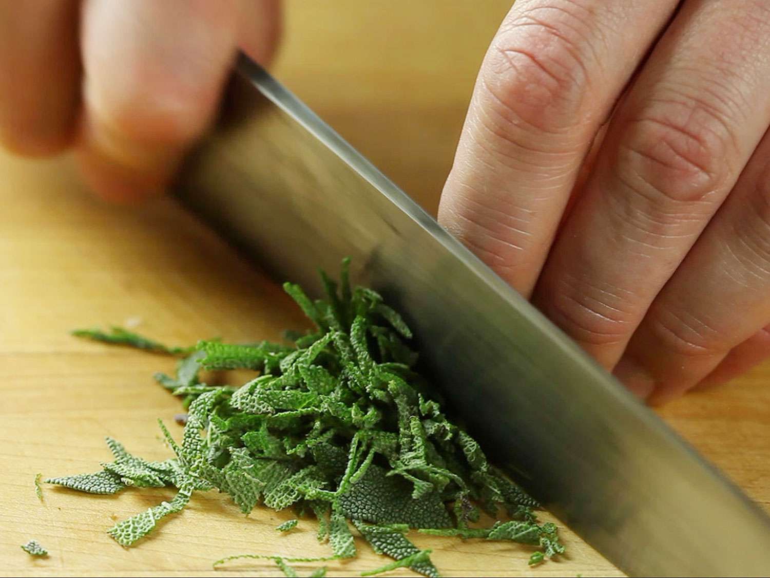 A knife slicing fresh sage leaves. There are also two hands in the image.