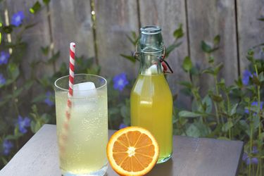 A glass of DIY orange soda with a striped straw next to a bottle of orange concentrate and a sliced orange.