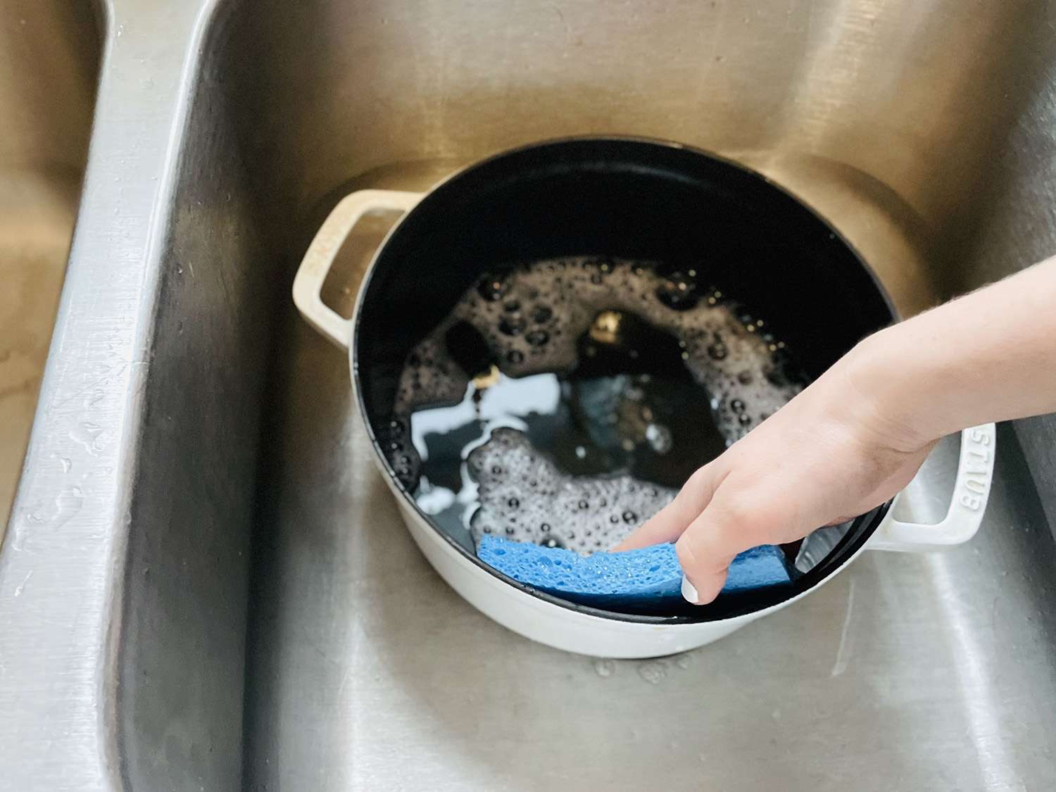 a hand using a sponge to clean a white dutch oven in the sink.
