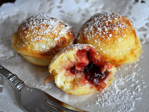 Three ebelskivers on a white cloth, dusted with powdered sugar. One is torn is cut in half, showing the lingonberry jam filling.