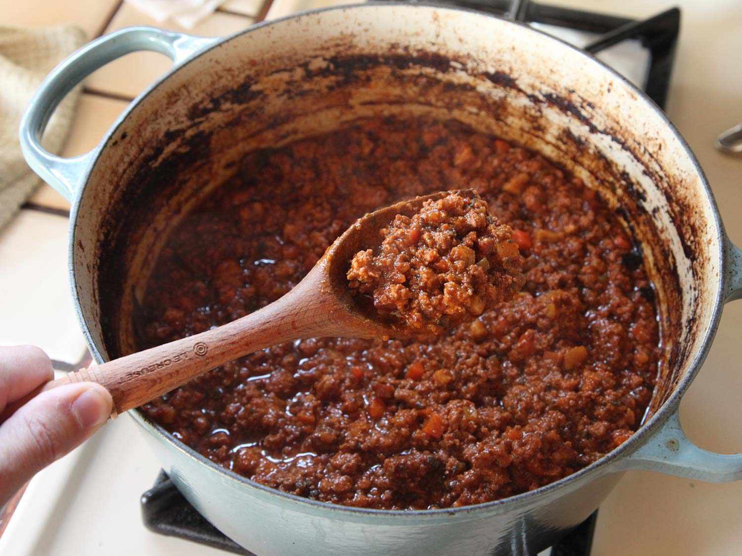 A wooden spoonful of meat sauce taken from a Dutch oven with the remainder of the sauce underneath. 