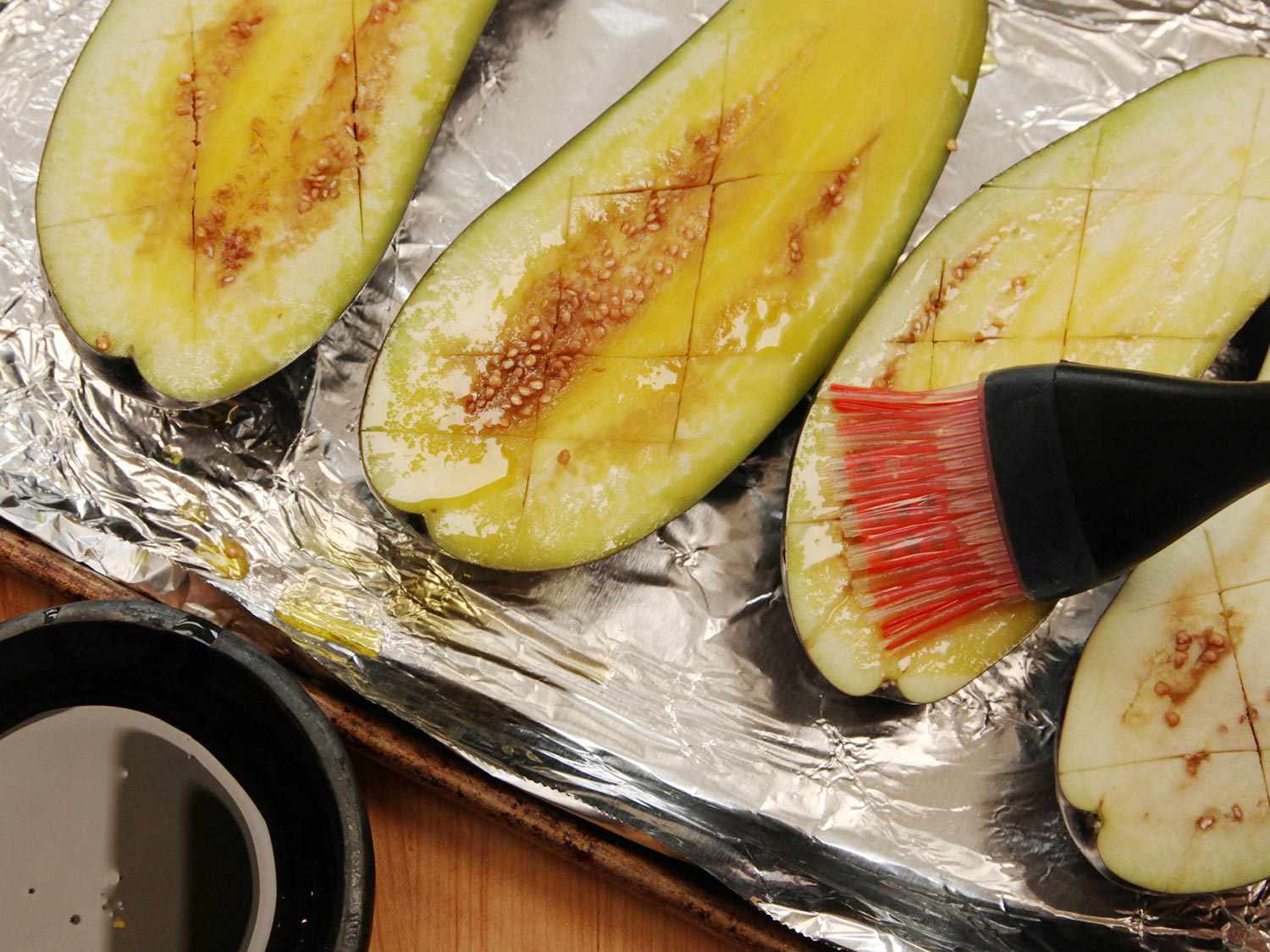 Halved eggplants are scored, arranged on a foil-lined sheet pan, and brushed with olive oil.