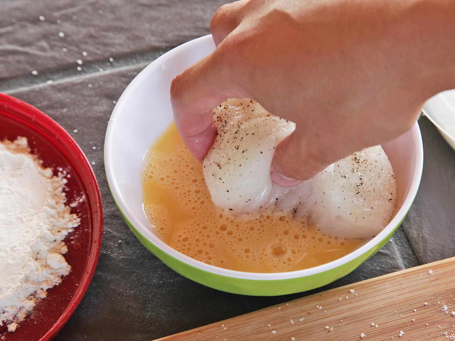 Author dips the floured side of a fish fillet portion into a bowl of beaten eggs.