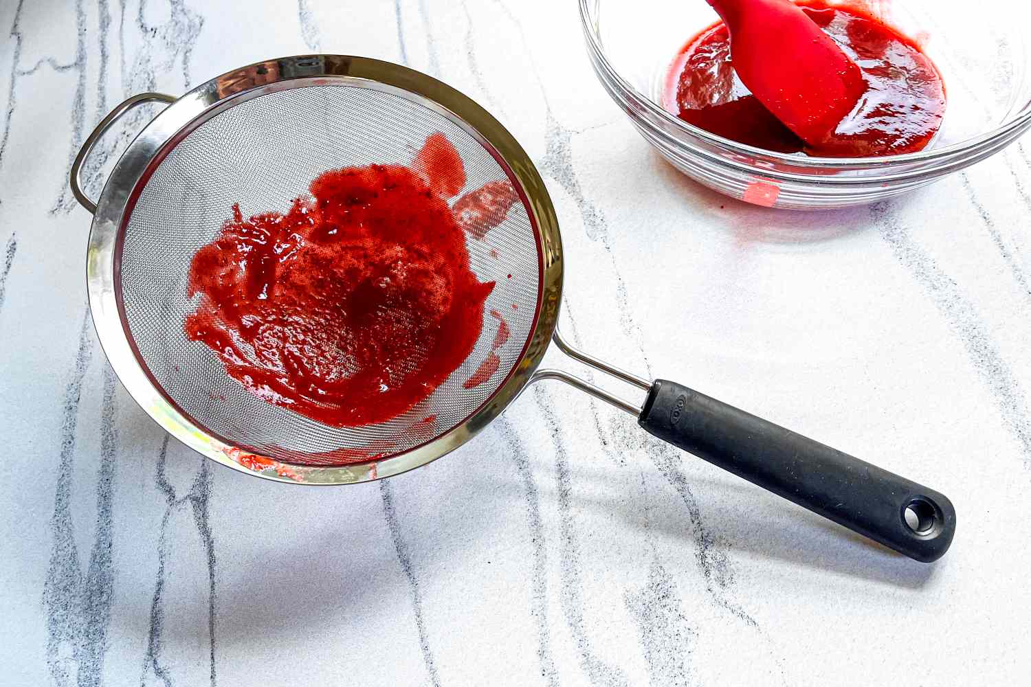 OXO Good Grips 8-Inch Double Rod Strainer with pressed fruit pulp beside a bowl containing a red liquid and a spatula