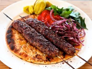 Two Adana kebabs on a plate with flatbread, sumac onions, parsley, tomatoes, and pickled peppers. 