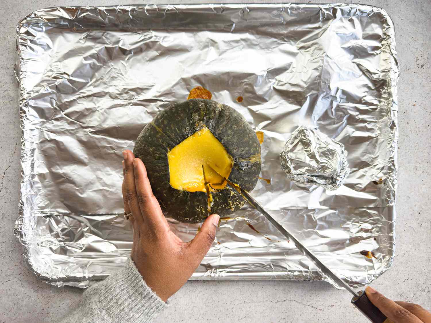 Hands cutting a slice of kabosha with a knife on a foil lined baking sheet 