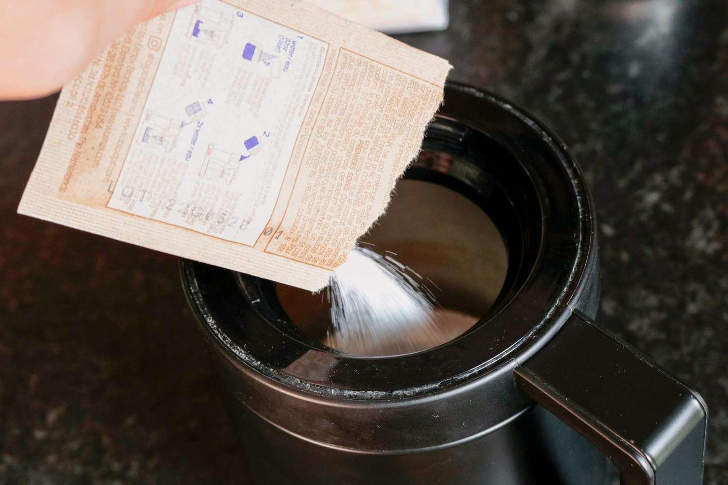 A packet of cleaning powder being emptied into a thermal coffee carafe