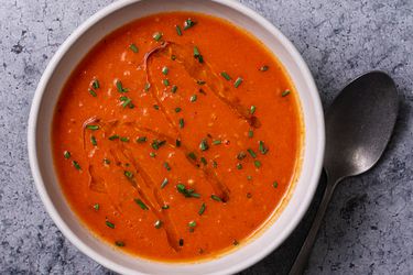 A bowl of creamy vegan tomato soup on a stone background with a spoon to the right of the bowl. The surface of the soup is drizzled with olive oil and sprinkled with chopped herbs.