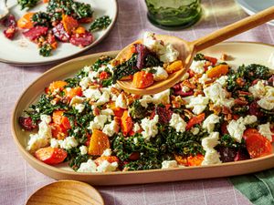 A platter containing kale salad with roasted root vegetables and crumbled cheese served with a wooden spoon