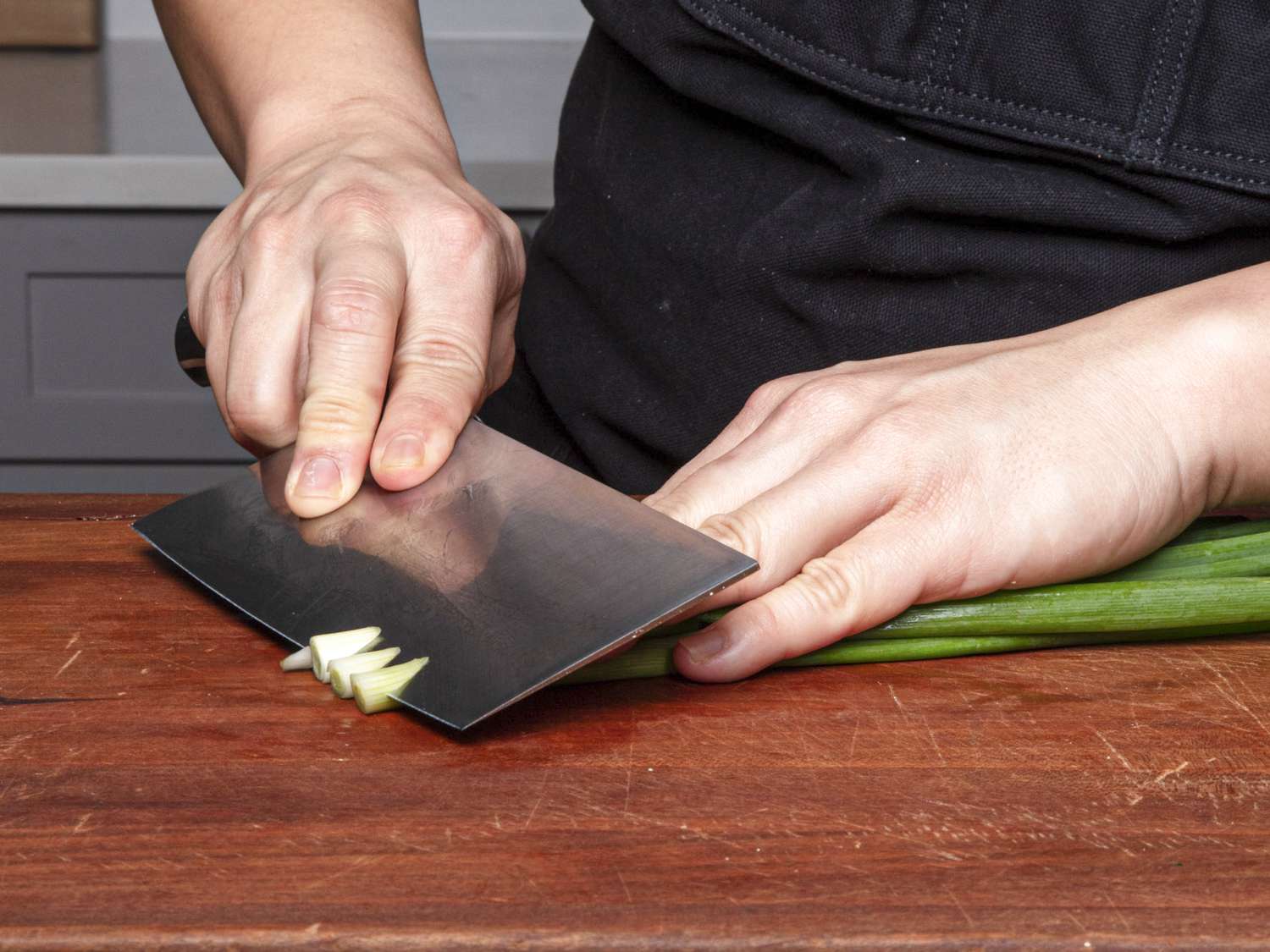 Scallions being cut using the reverse bias, where the fingers of the non-dominant hand lay on top of the ingredient and form the incline that will determine the cut; the knife blade is perpendicular to the cutting board, and angled slightly down.