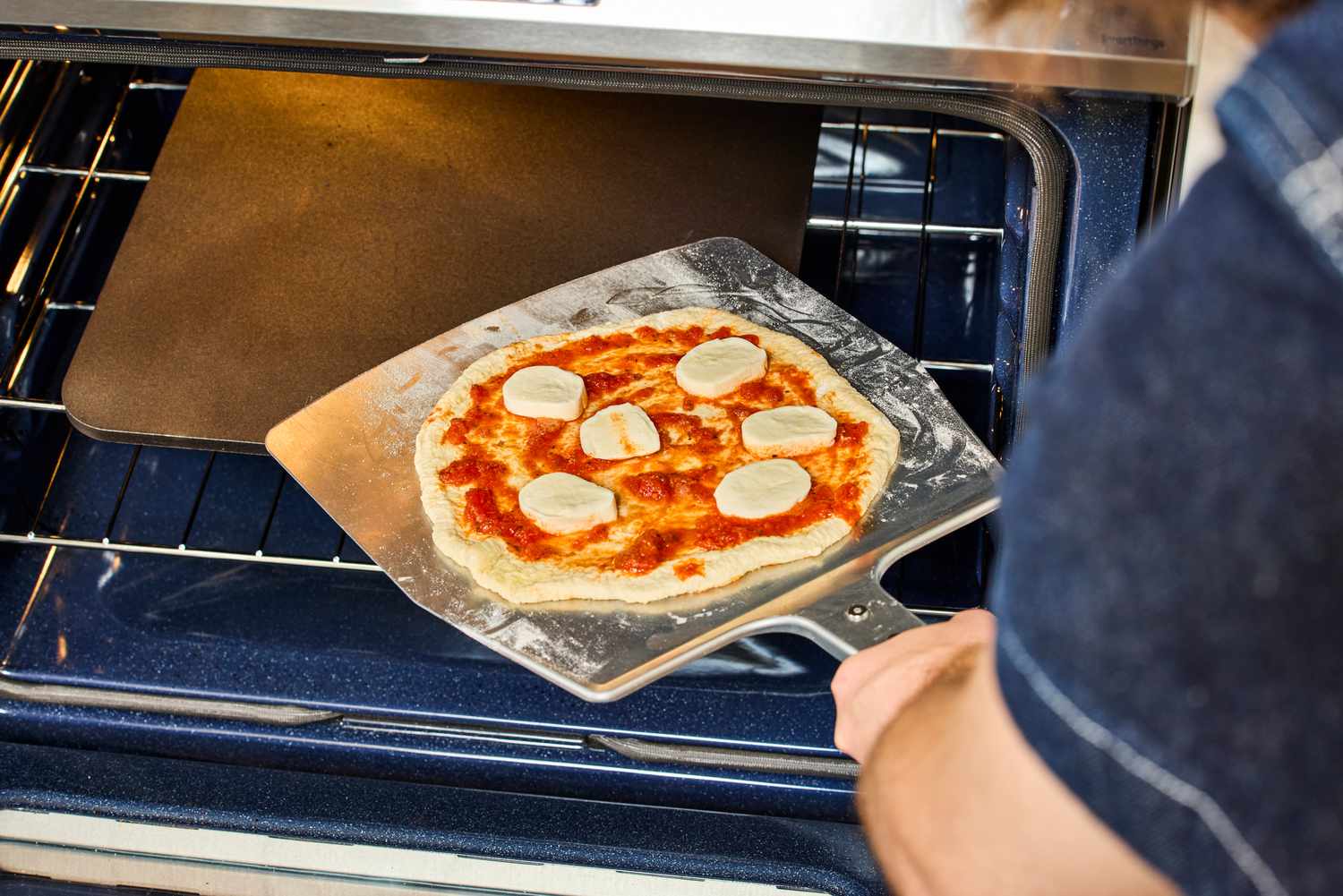 A person adding a pizza to a pizza steel in the oven.