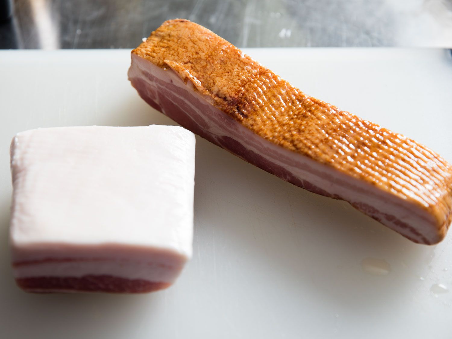 Slab of salt pork and a slab of bacon next to each other on a white cutting board.