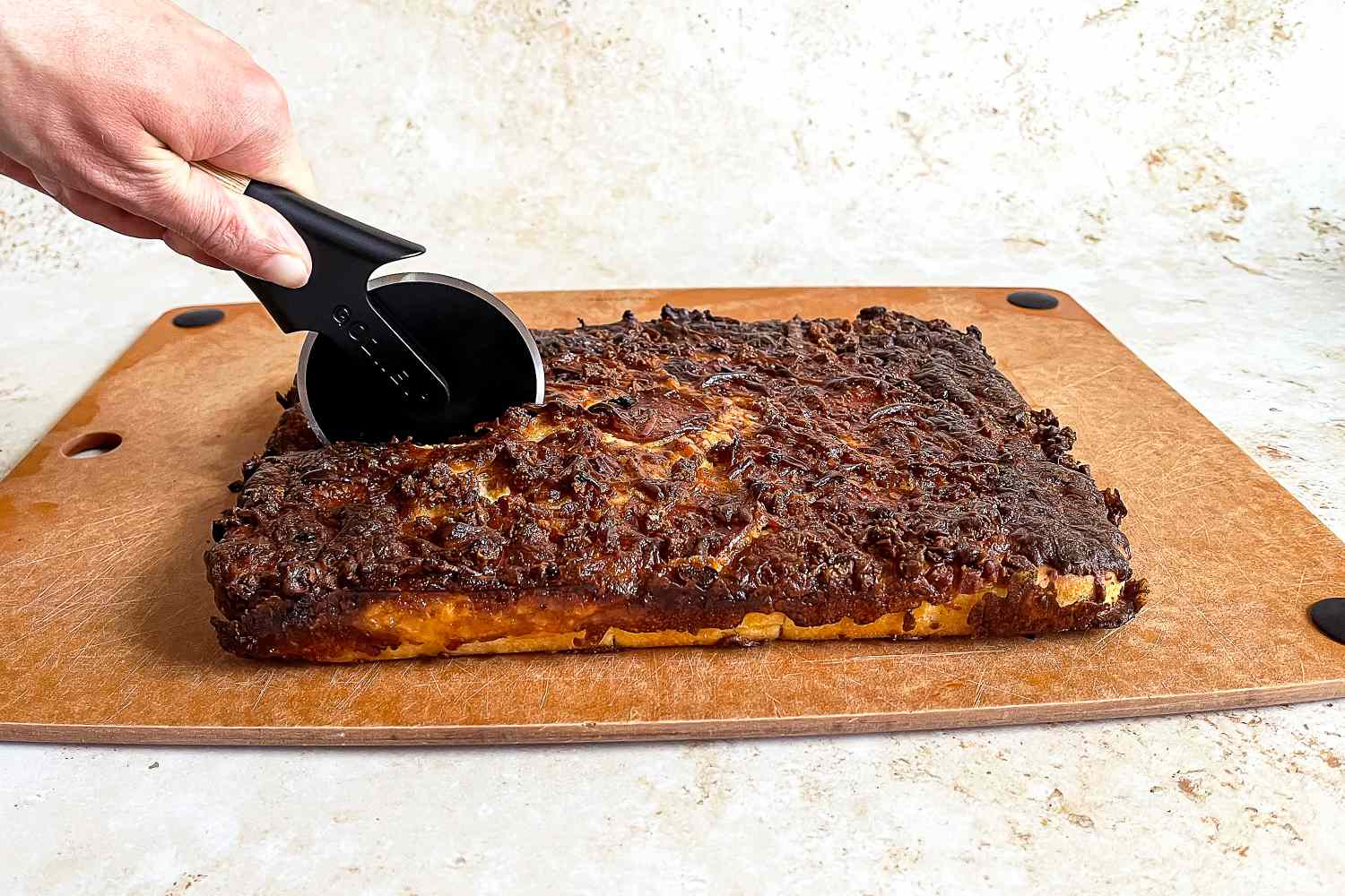 A hand using a Gozney pizza cutter on a rectangular baked dish on a wooden surface