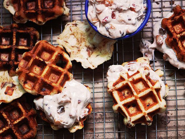 Overhead view of Cinnamon Roll Waffles on a cooling rack over a rimmed baking sheet. A ramekin of Bacon-Apple Frosting is in the frame and one of the waffles has been frosted.