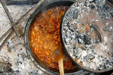 A Dutch oven filled with chili cooking over campfire coals and being stirred with a wooden spoon.