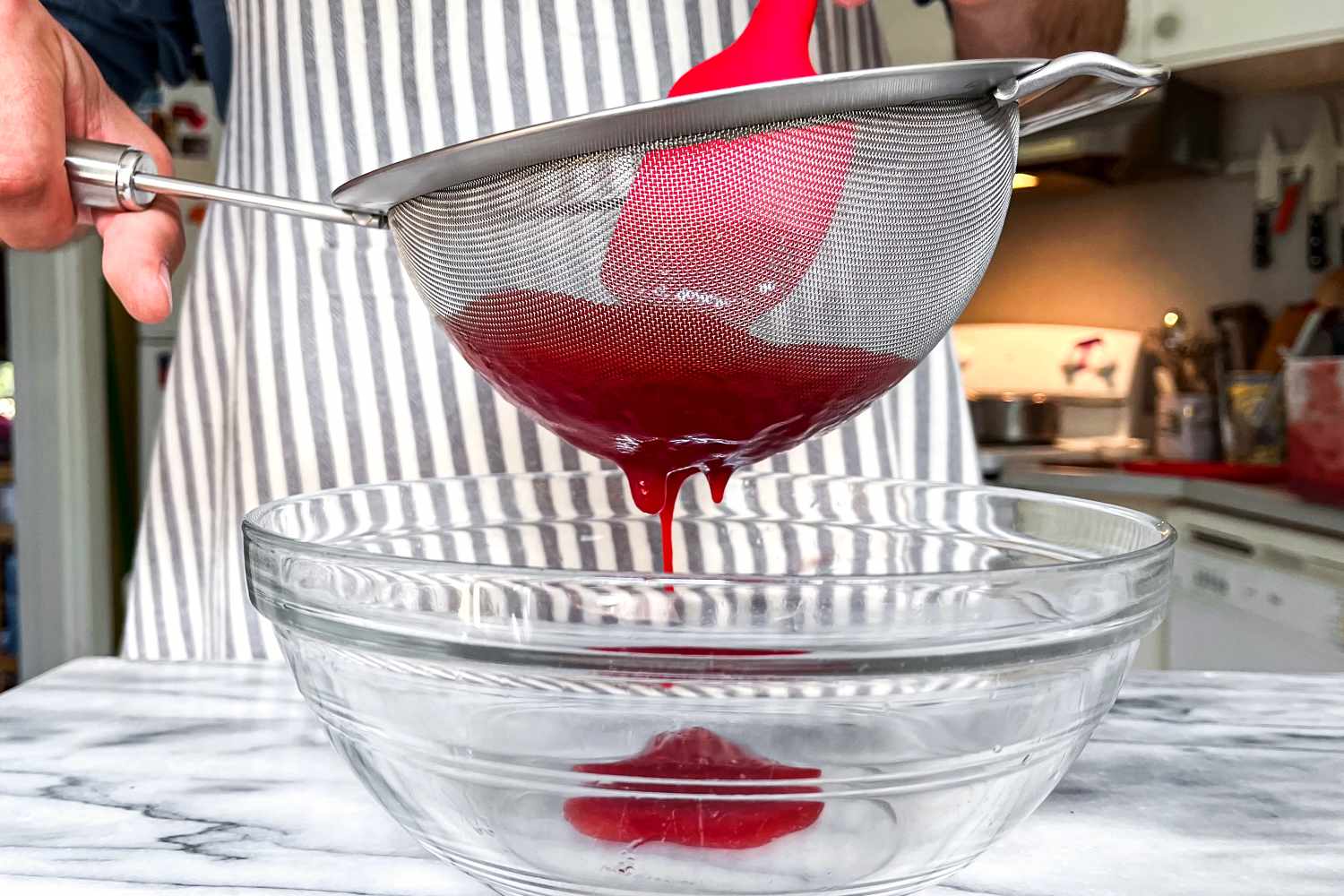 A person strains a red liquid through the Rösle Stainless Steel Round Handle Kitchen Strainer into a glass bowl using a spatula in a kitchen setting
