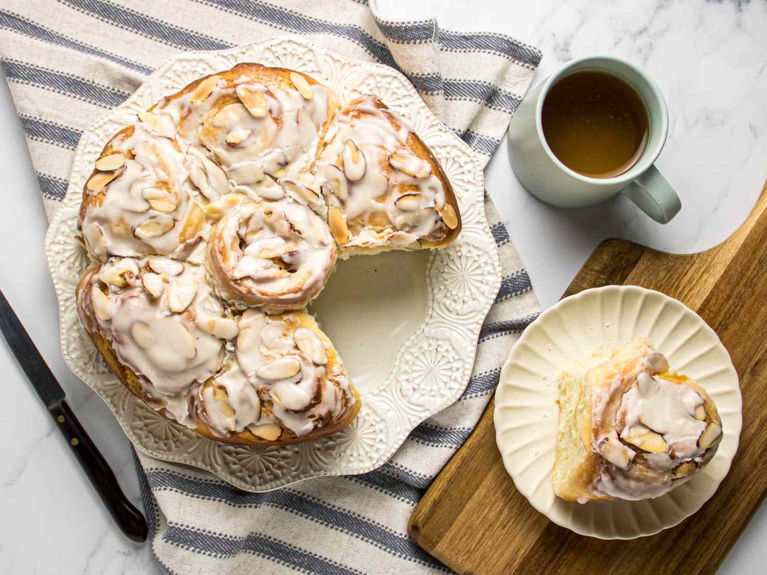 almond frangipane roll on white plate on wooden board, with large plate of rolls, coffee, and a striped napkin. 