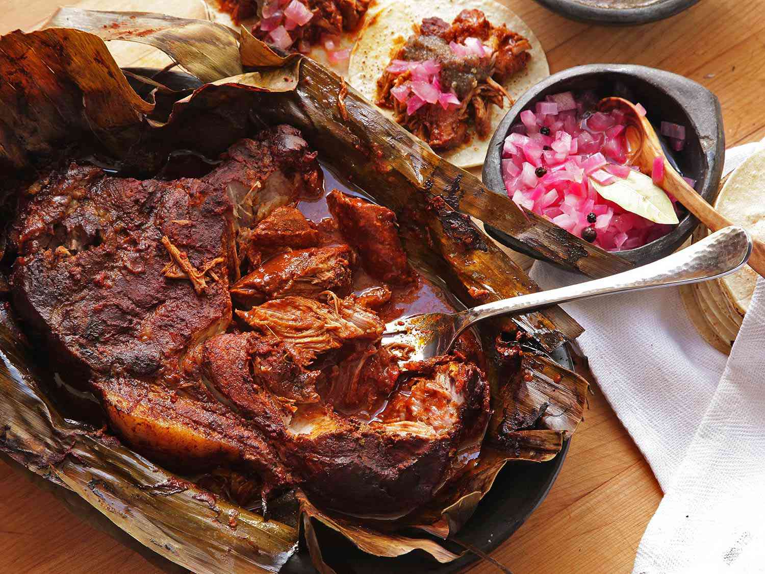 Overhead view of cochinita pibil, partially unwrapped in its banana-leaf cocoon. A patch of the pork roast has been shredded with a fork.