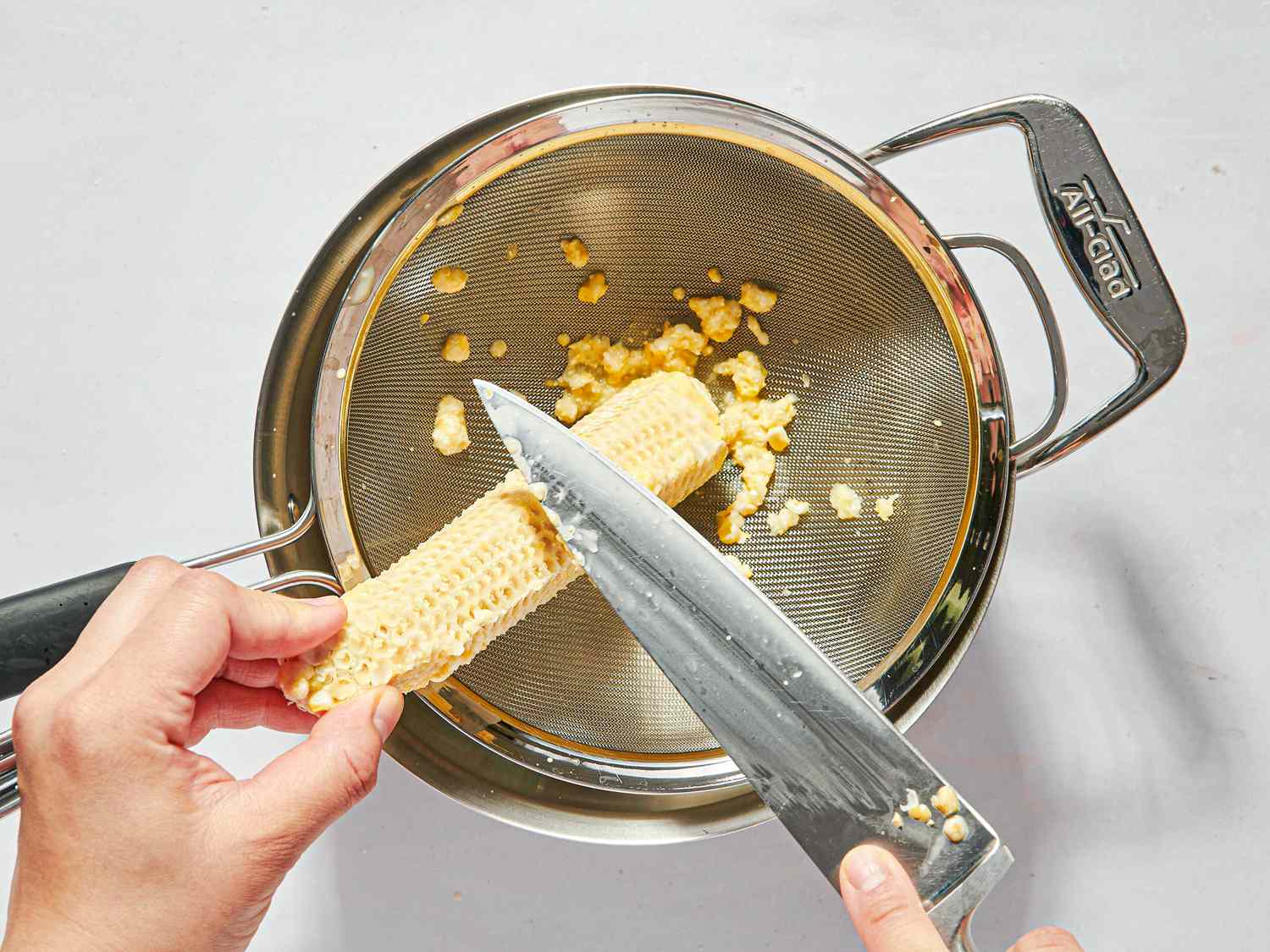 Hands cutting corn from a cob over a metal strainer with a knife