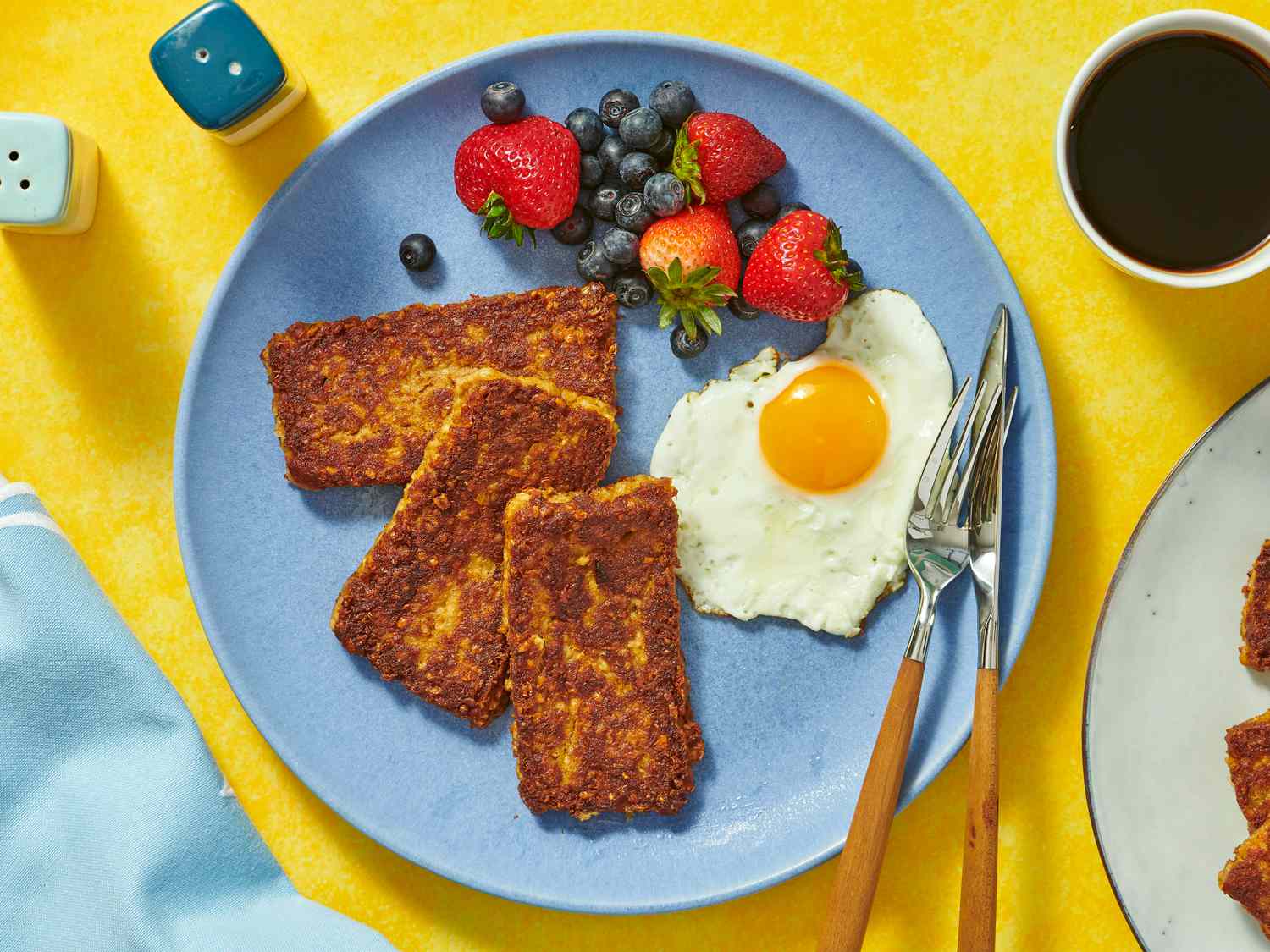 overhead angle of goetta, berries and fried egg on a blue plate and yellow background. coffee and plate of goetta in the background