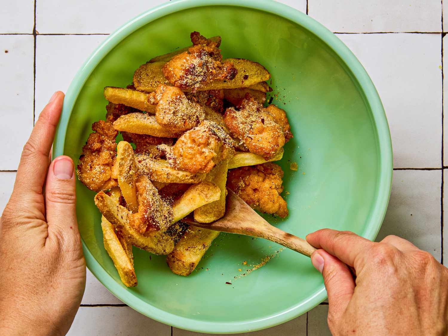 A bowl of fried food including fries and breaded pieces being mixed with a wooden spoon by hands