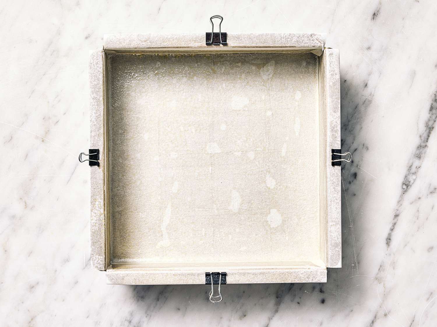 Square baking pan prepared with parchment paper and binder clips on a marble surface
