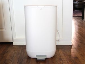 A white trash bin in a room with wooden flooring and a partial view of a door and furniture in the background