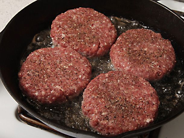 Four burger patties are pan-fried in a cast iron skillet.