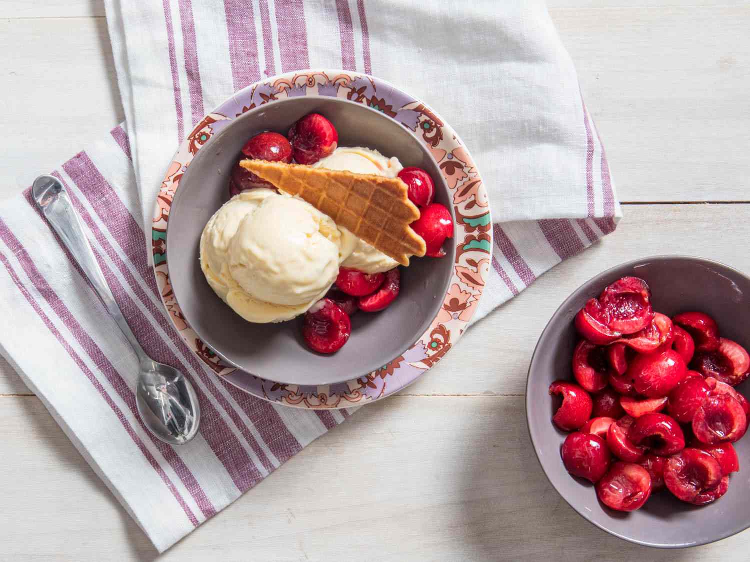 Overhead view of a triple-scoop bowl of no-churn mascarpone ice cream topped with halved, macerated cherries and a waffle-cone tuille. A bowl of the halved cherries sits off to one side.
