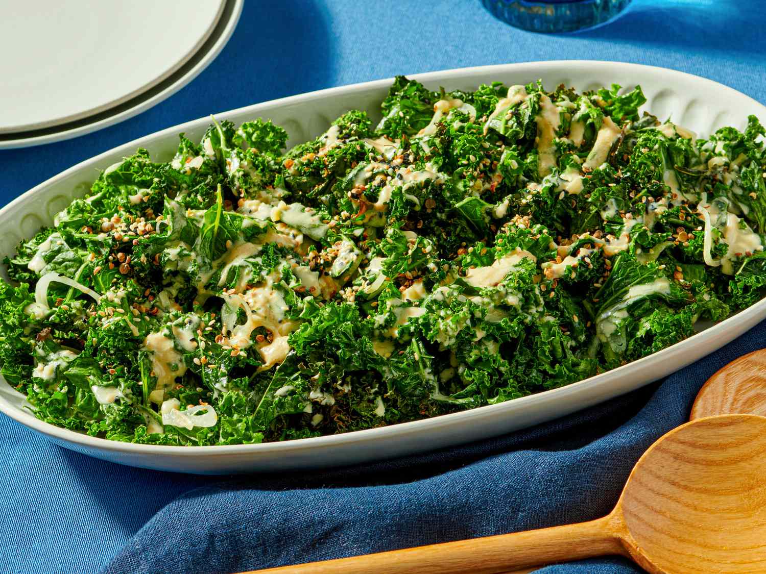 A dish containing roasted kale salad with toppings on a blue tablecloth wooden serving utensils on the side